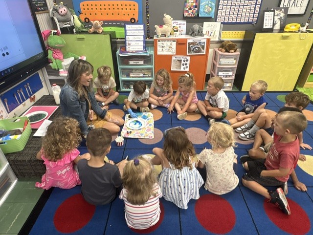 A teacher and a circle of young students watch a science demonstration together on the classroom rug