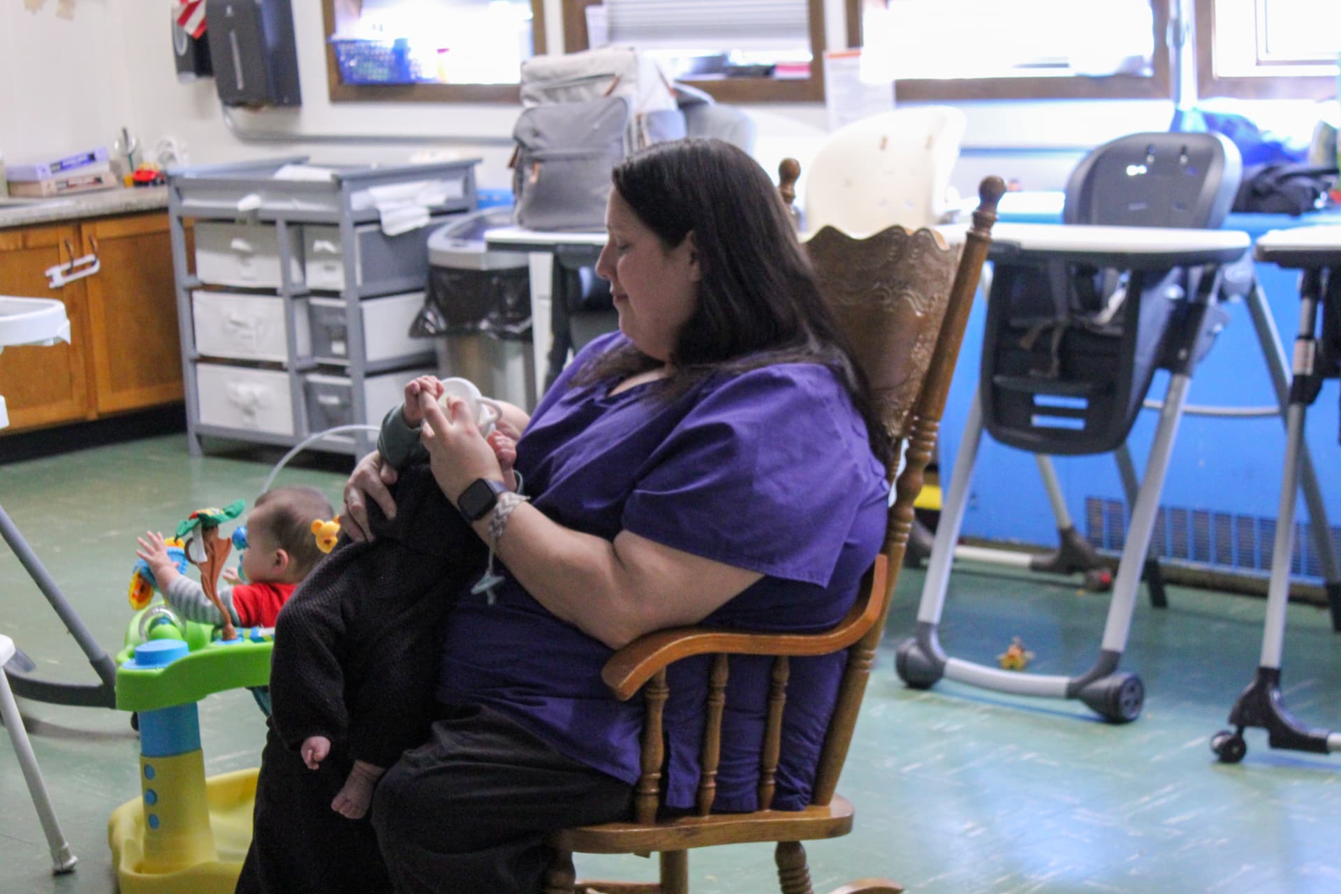 A caregiver in purple scrubs sits in a wooden rocking chair holding an infant while a toddler plays in an activity saucer nearby