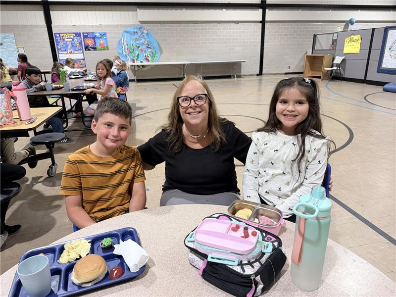 A teacher sits between two students at a cafeteria table with lunch trays in front of them