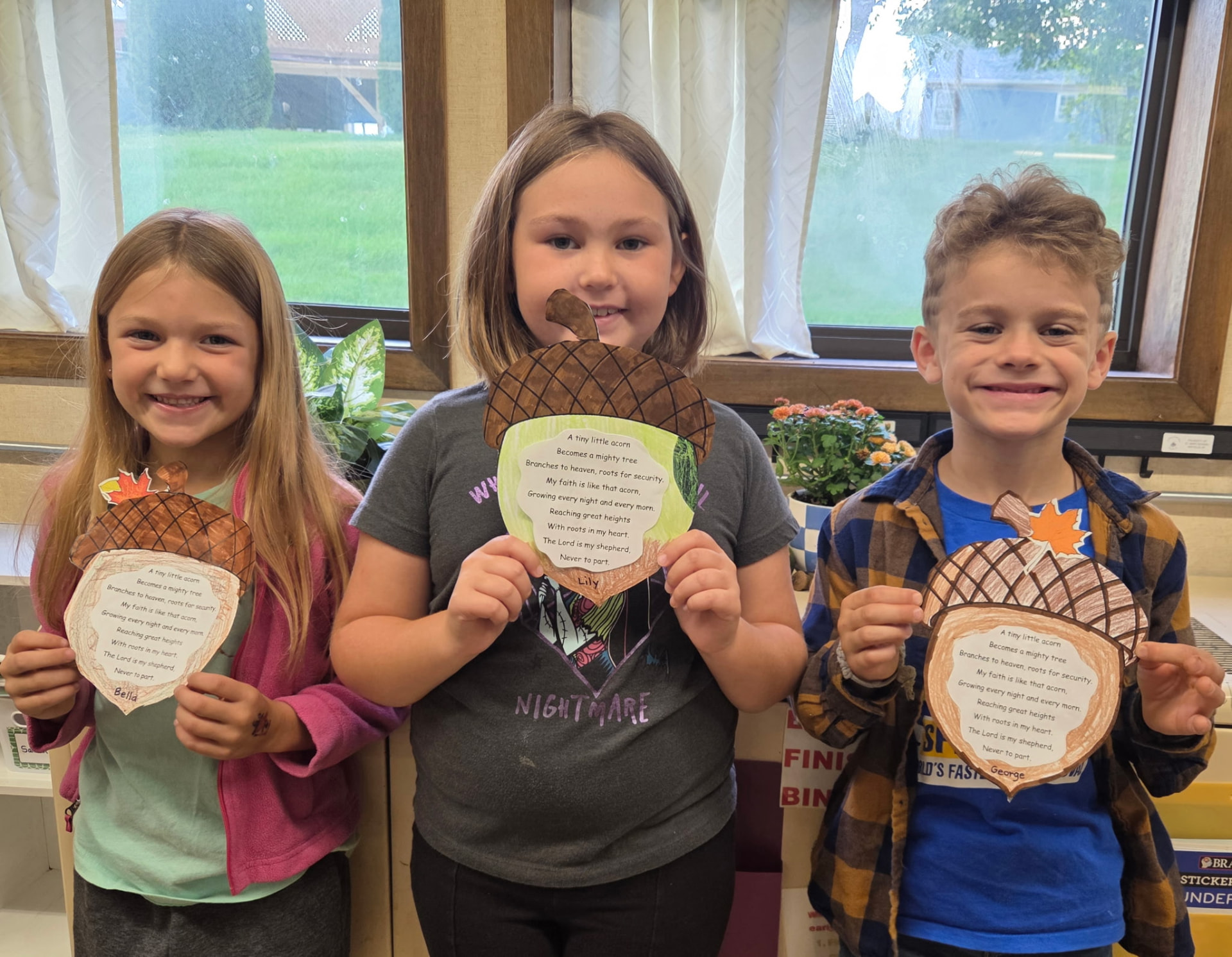 Three children stand at a classroom window holding up paper acorn crafts with their names written on them