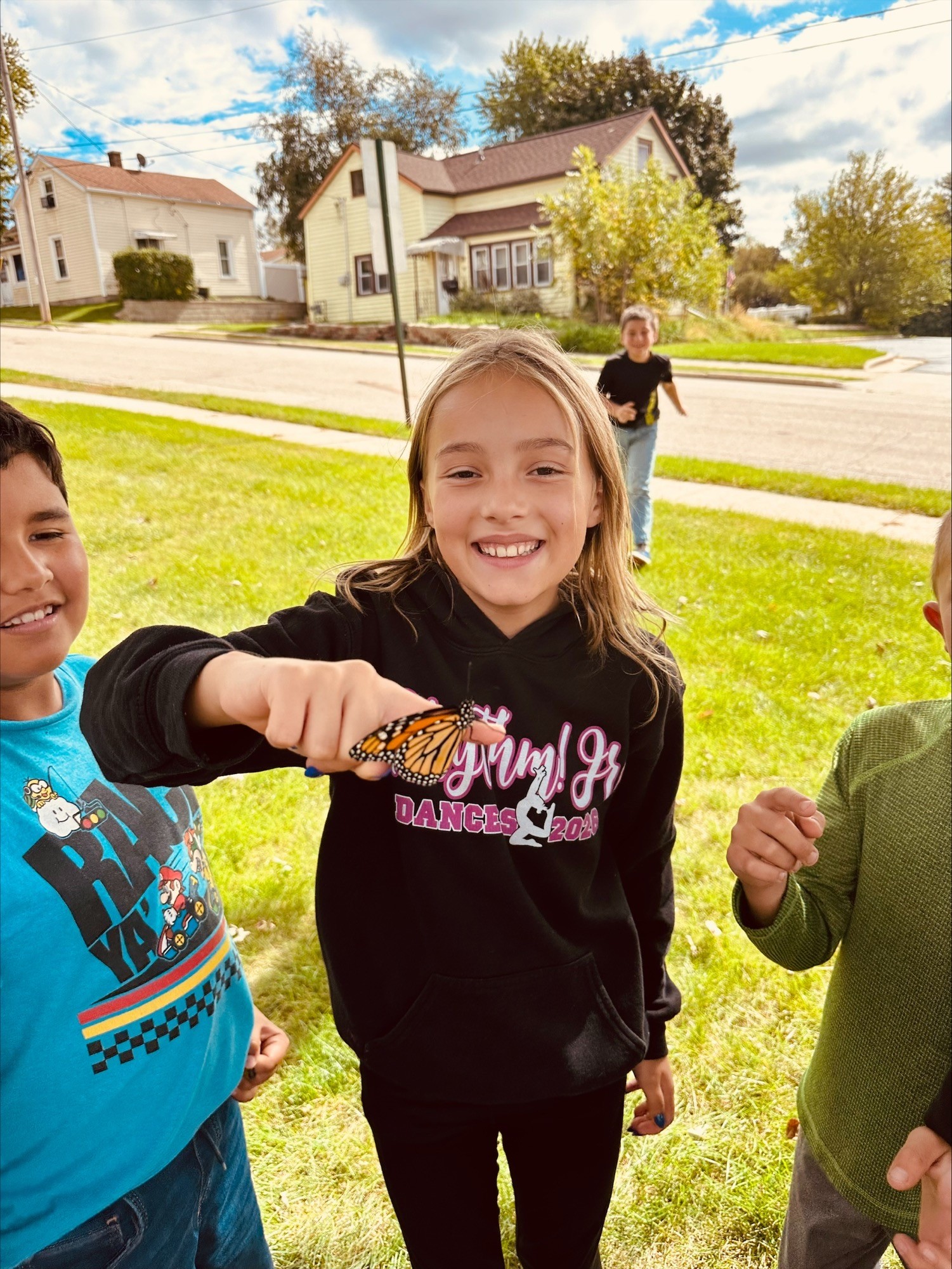 A smiling girl holds an orange and black monarch butterfly on her fingertip while classmates gather on the lawn behind her